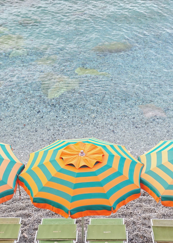 Luftaufnahme eines orange-grün gestreiften Sonnenschirms am Kiesstrand mit türkisem Wasser