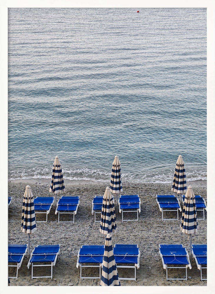 Blaue Strandliegen und geschlossene gestreifte Sonnenschirme am Kiesstrand vor dem Meer
