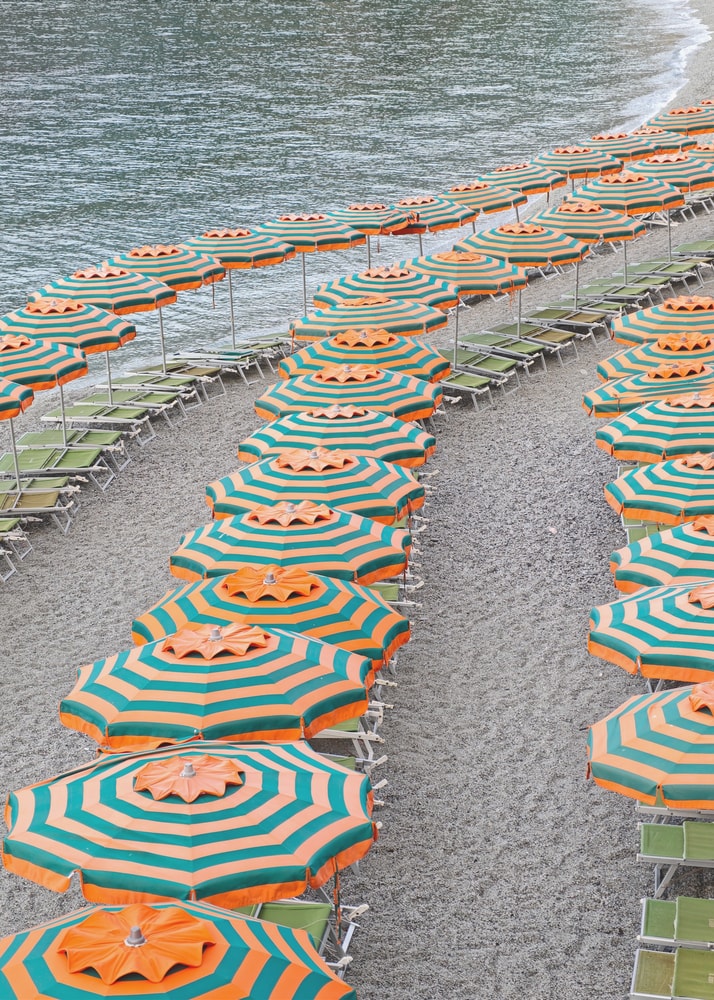 Luftaufnahme geschwungener Reihen orange-türkiser Sonnenschirme an einem Kiesstrand