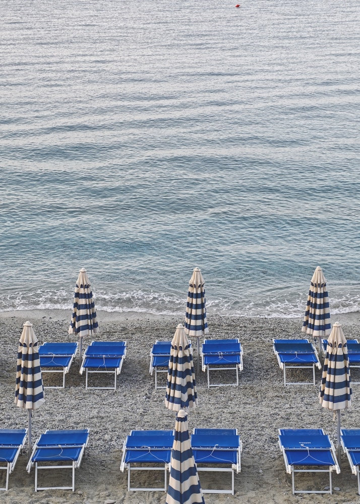 Blaue Strandliegen und geschlossene gestreifte Sonnenschirme am Kiesstrand vor dem Meer