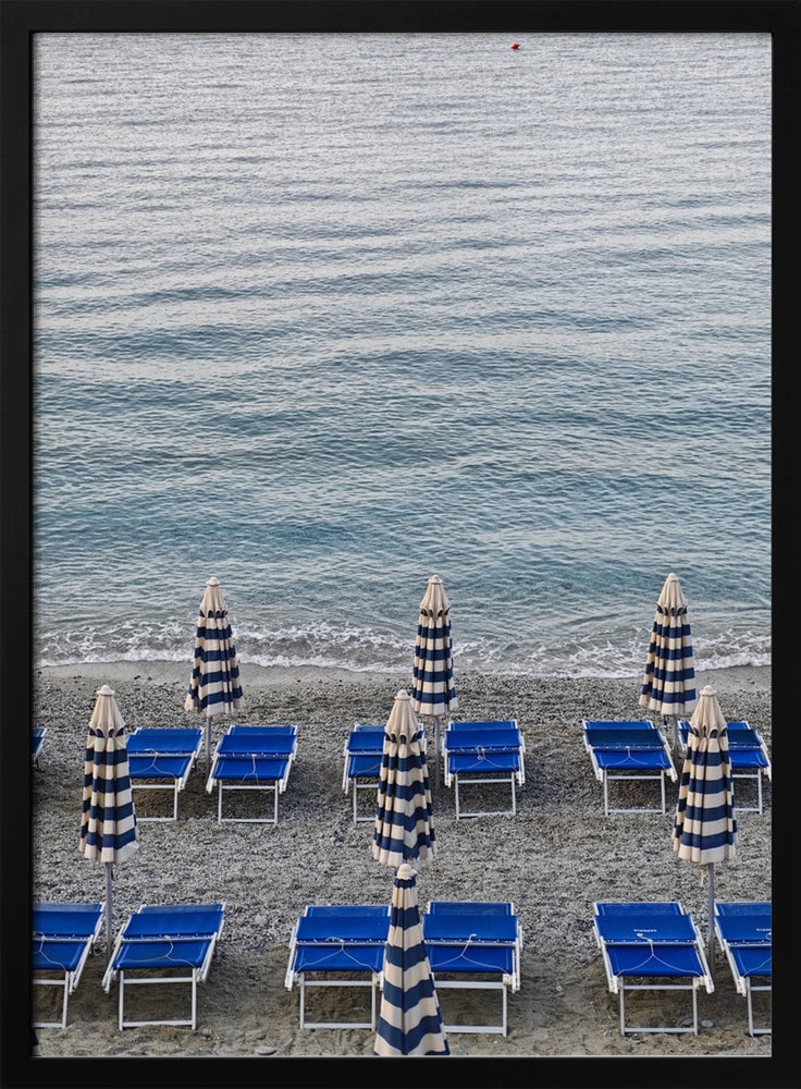 Blaue Strandliegen und geschlossene gestreifte Sonnenschirme am Kiesstrand vor dem Meer